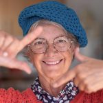 Enevjecimiento de los dientes. Retrato de una hermosa anciana sonriendo con anteojos y gorra azul gesticulando con las manos